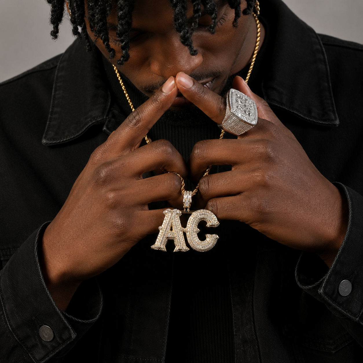 Person wearing a gold chain with a 'AC' pendant and a large ring, against a dark background.