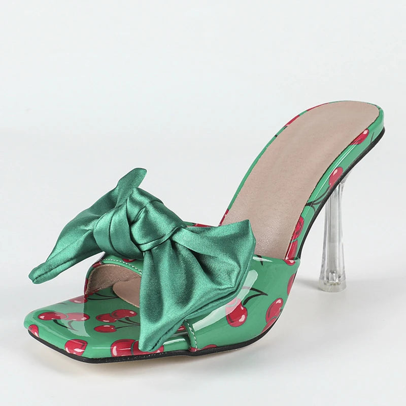 Green high-heeled shoe with a large bow and cherry pattern on a white background