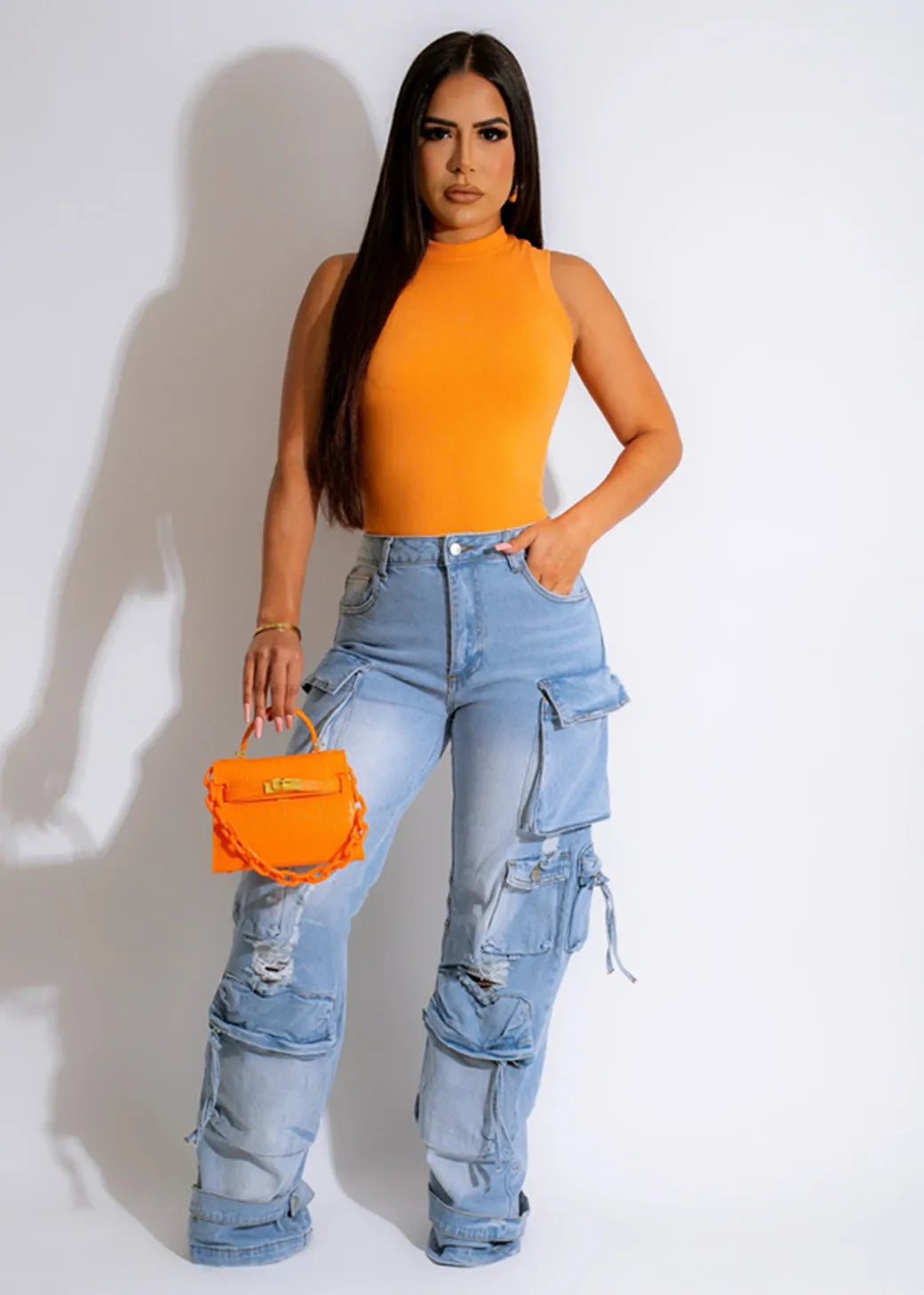 Woman wearing an orange top and blue jeans, holding an orange handbag against a white background