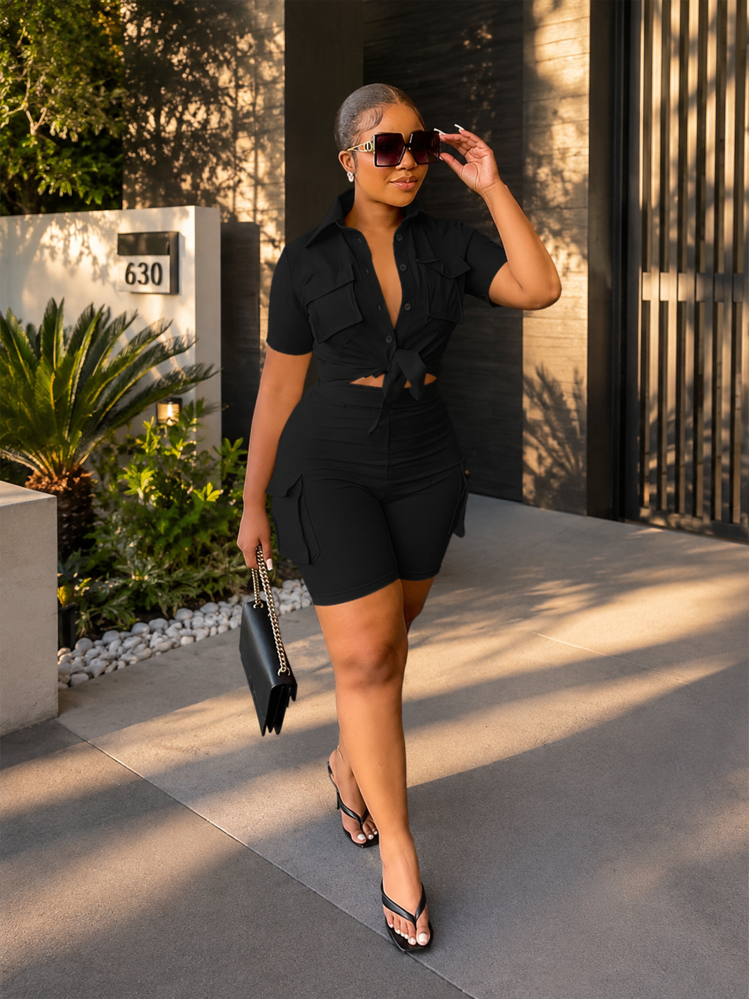 Woman in a black outfit standing outdoors near a building with plants.
