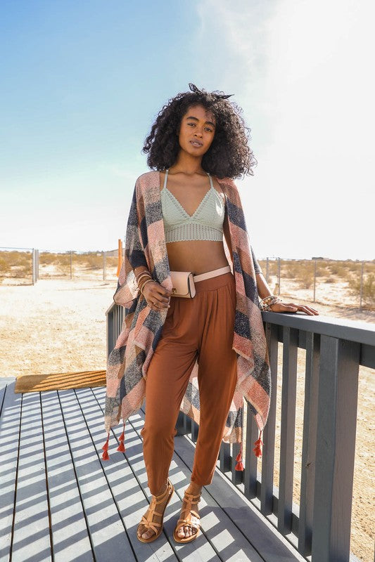 A woman standing on a wooden deck wearing a multicolored buffalo checkered kimono, paired with brown trousers and a green top, accessorized with a pink belt and sandals.